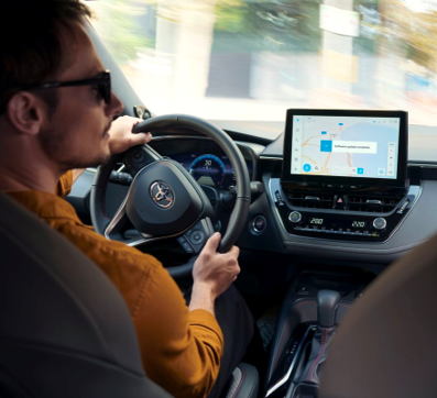 A man in an orange jumper turns rights as he drives a Toyota car. The car's multimedia system displays a software update.